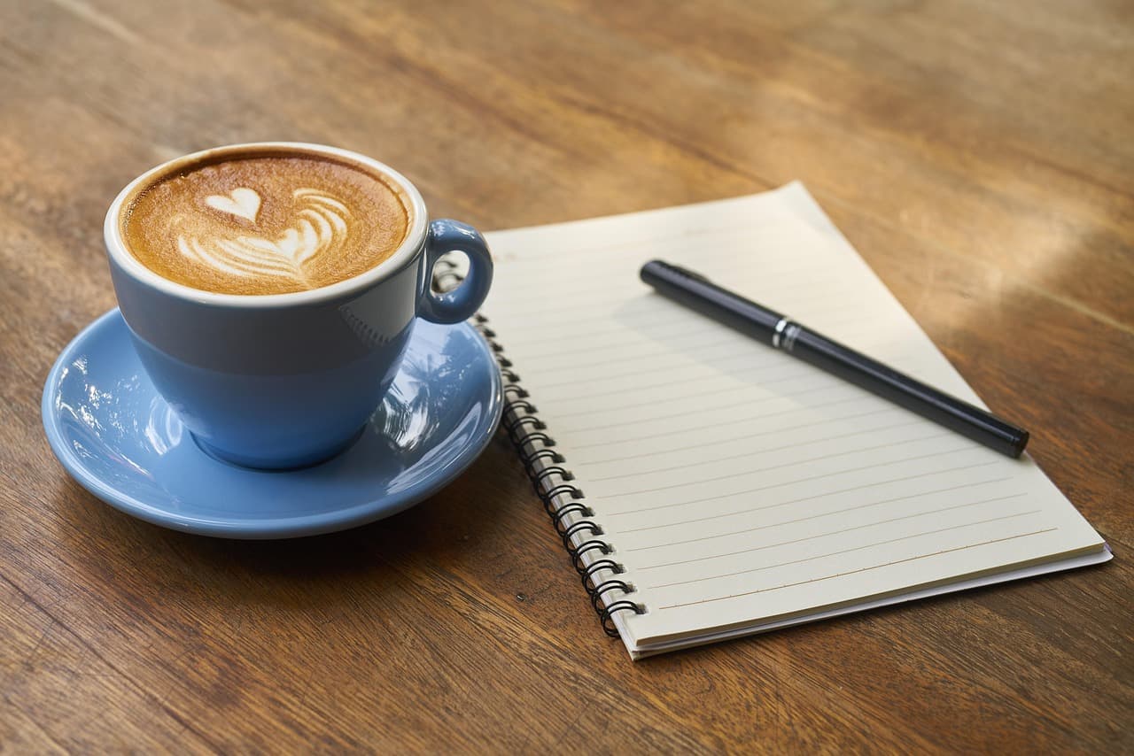 A latte with heart latte art beside a notebook on a wooden table